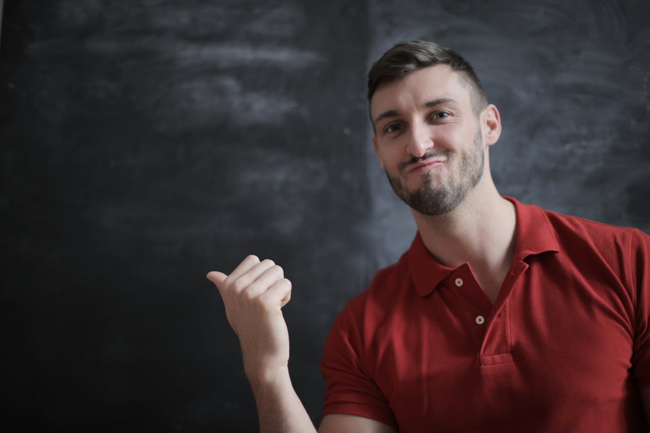 Portrait of a young man in a red polo, gesturing confidently indoors with a chalkboard background.