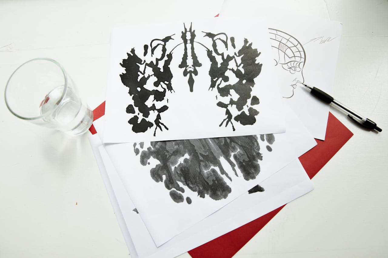 An artistic arrangement of inkblot test sheets with a pen and glass on a table.